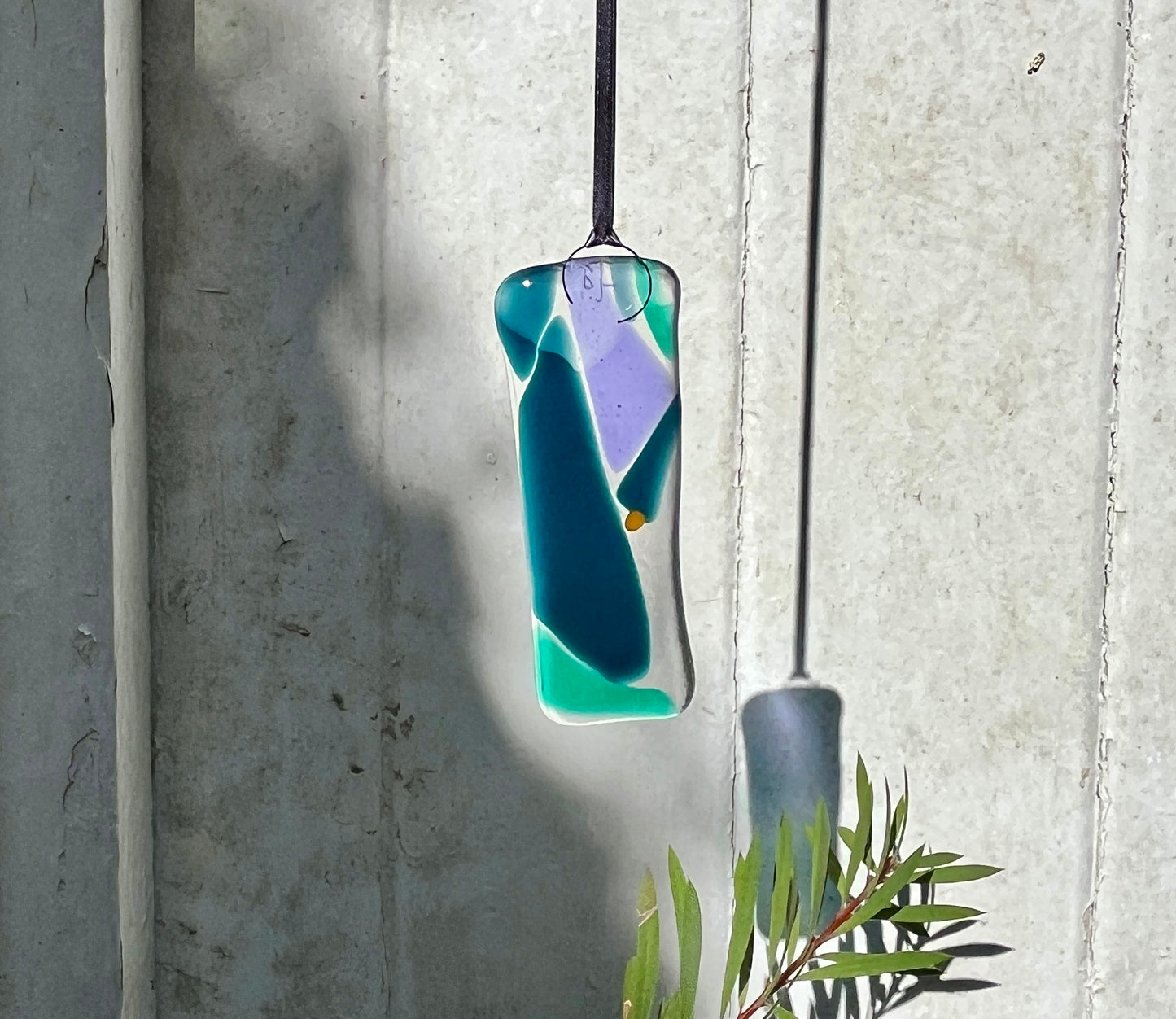 Glass Bryony Lane pendant with blue and green colors hanging against a concrete wall.