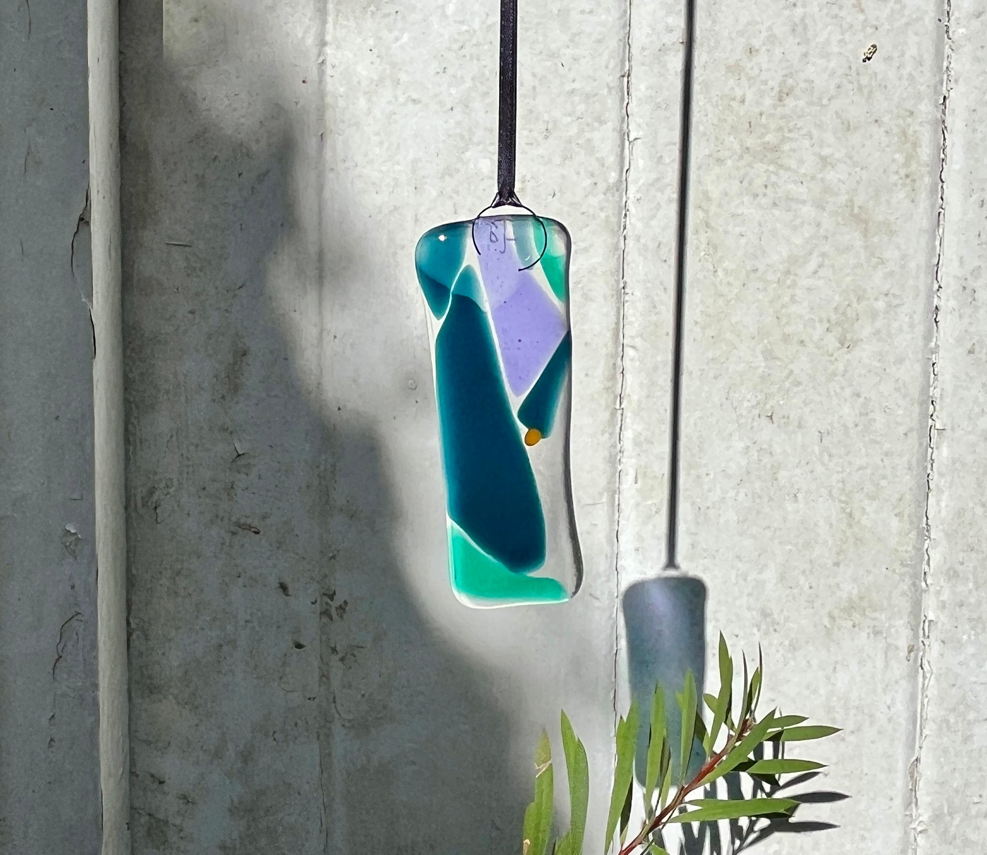Glass Bryony Lane pendant with blue and green colors hanging against a concrete wall.