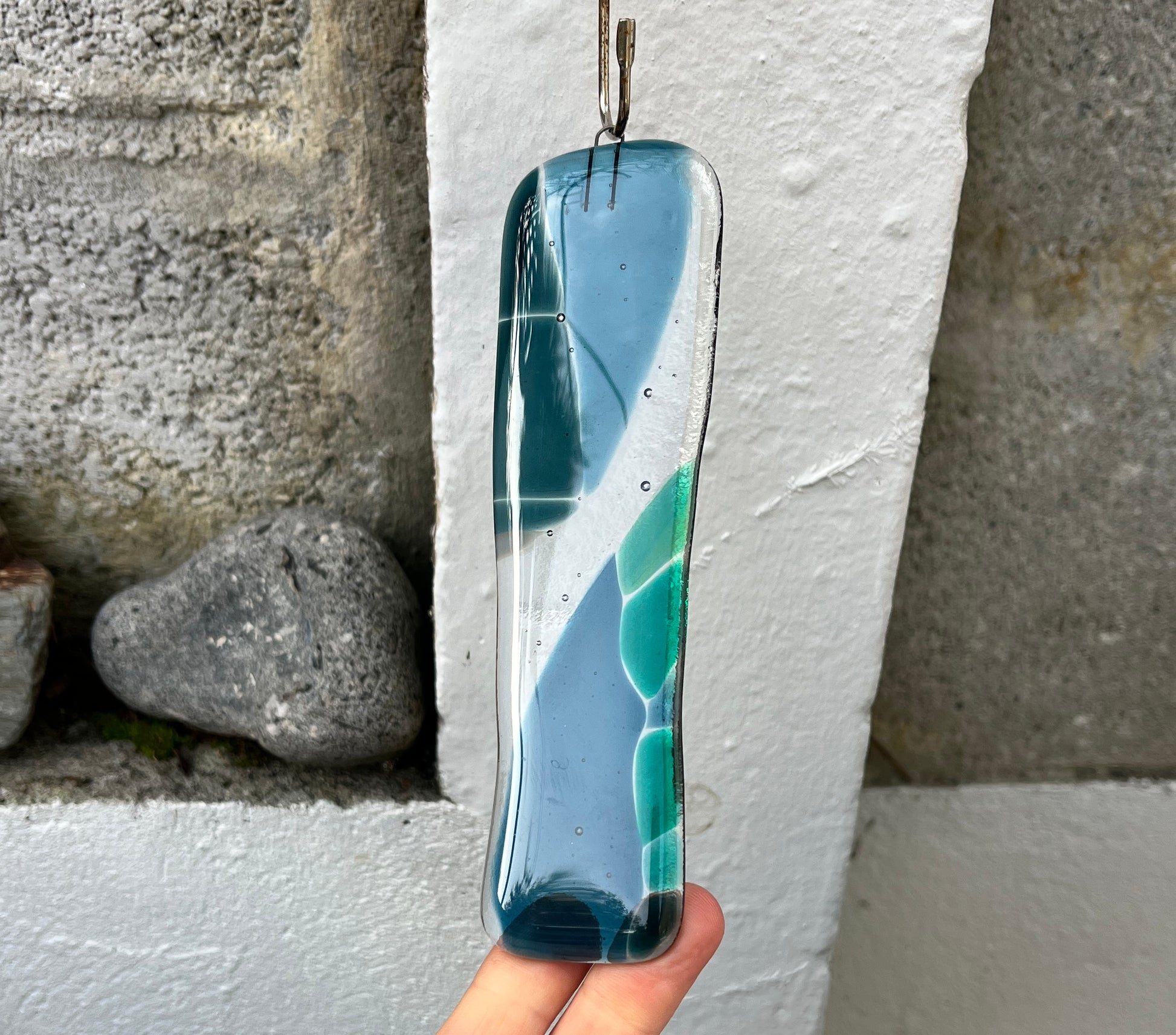 Glass pendant with blue and green design held against a stone wall.